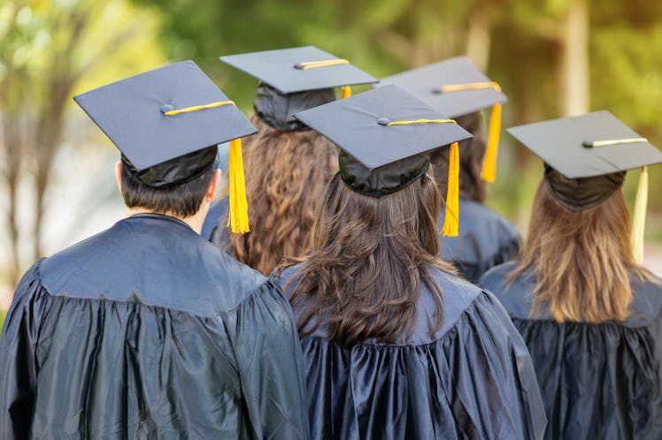 Diverse college graduates listen to speaker during graduation. They are wearing black caps and gowns...