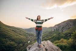 Young female hiker standing on a rock high on mountain in sunset. Your May 10 zodiac sign daily horo...