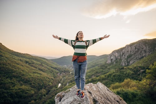 Young female hiker standing on a rock high on mountain in sunset. Your May 10 zodiac sign daily horo…
