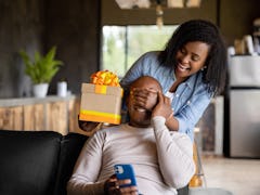 African American woman at home surprising her husband with a gift and covering his eyes