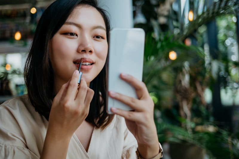 Image of a young Chinese woman putting on lipstick using smartphone as a mirror