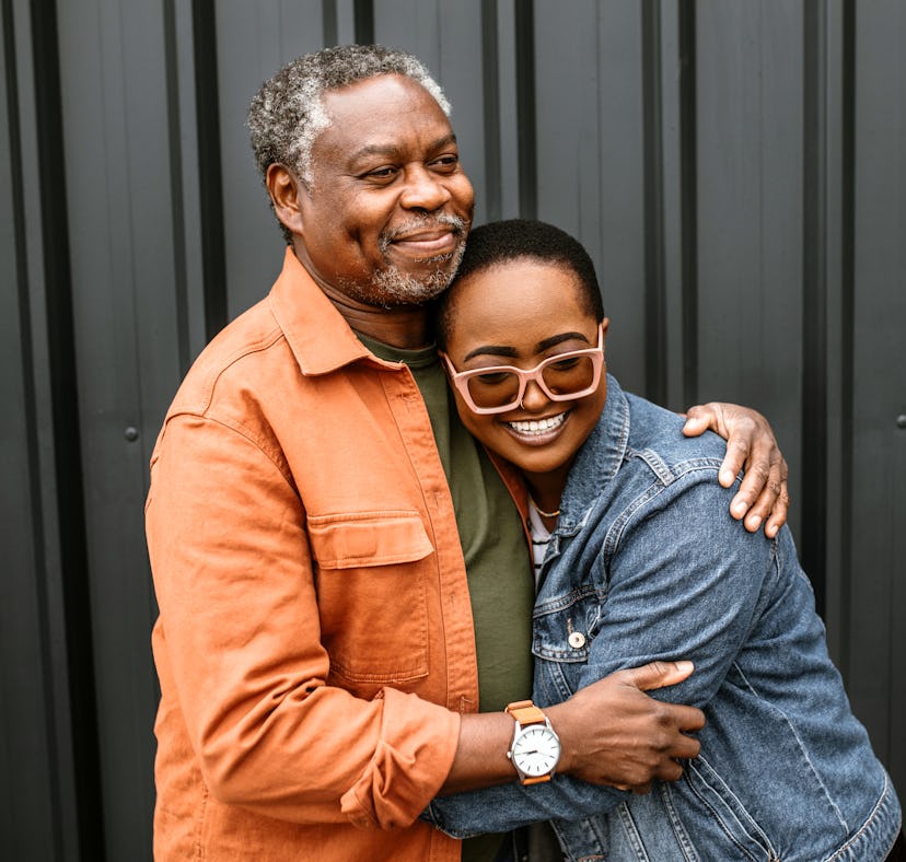 Portrait of a senior father and adult daughter standing in a front of a black door, smiling, embraci...