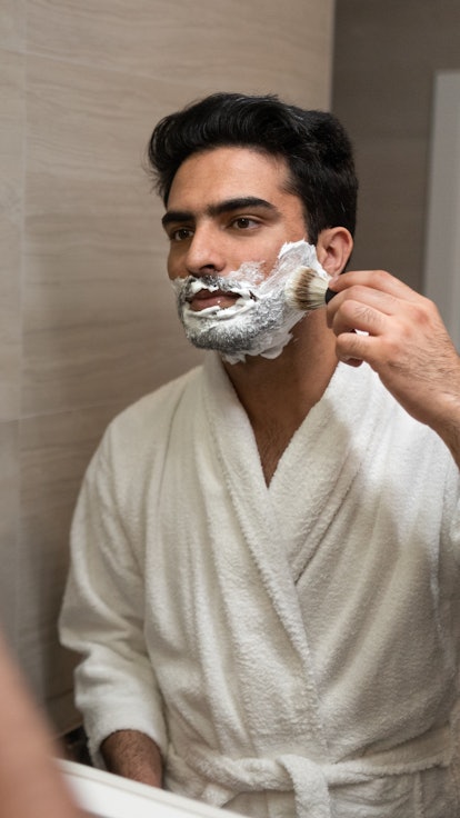 Young handsome muslim man standing in front of mirror in bathroom and shaving his beard.
