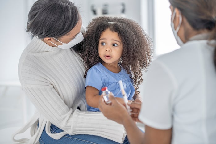 A female doctor holds out a vaccine vial and syringe as she talks with a young mother about a vaccin...
