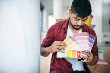 Young man sitting on the floor at his apartment, holding color swatch and choosing  color to paint h...