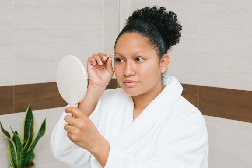 Back view of focused female in bathrobe plucking eyebrows while standing in bathroom looking into a ...