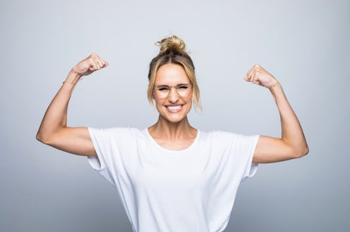Powerful beautiful woman wearing white top flexing muscle while standing against gray background.