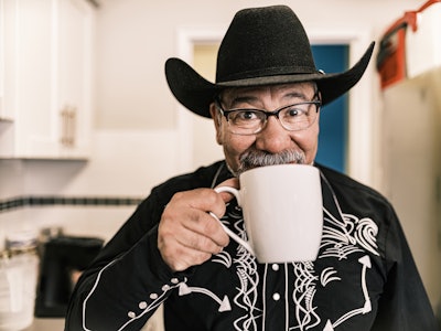 Mature Latin man dressed as Mexican Cowboy having a coffee. He has grey hair beard, wearing eyeglass...