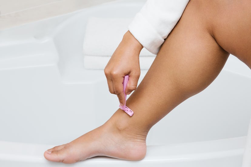 Crop unrecognizable female in bathrobe shaving legs with razor while doing skin care routine in bath...