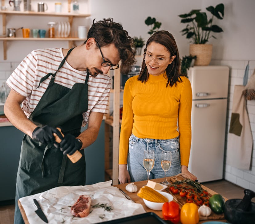 Young woman looking at boyfriend sprinkling spices on steak while standing in kitchen at home on Jul...