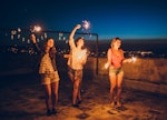 Three female friends dancing with sparklers on a rooftop on July 4, 2022 after reading their horosco...