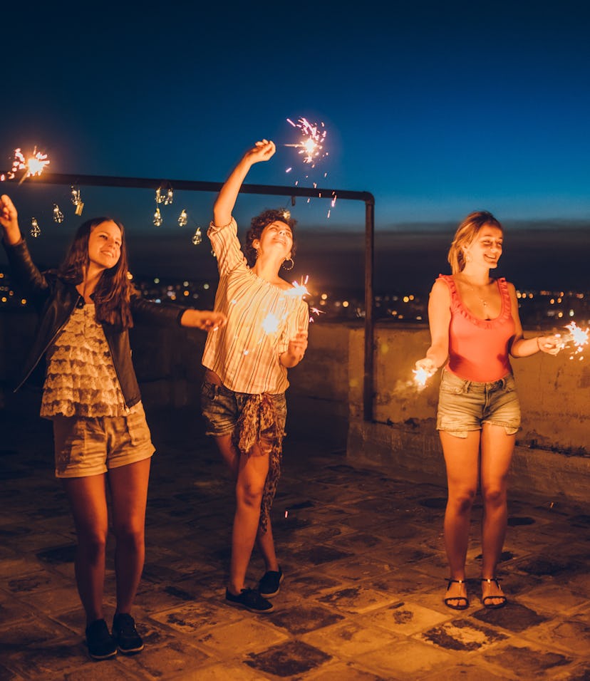 Three female friends dancing with sparklers on a rooftop on July 4, 2022 after reading their horosco...
