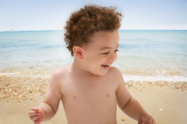 Toddler boy with curly brown hair smiling and looking to the side on a sandy beach with light blue w...