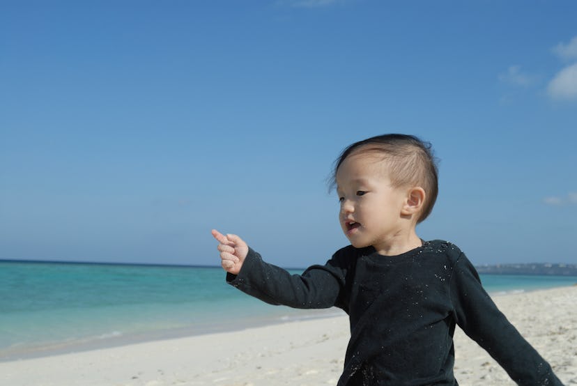 Toddler with dark hair on the beach in a black, long-sleeved onesie with arm extended, pointing fing...