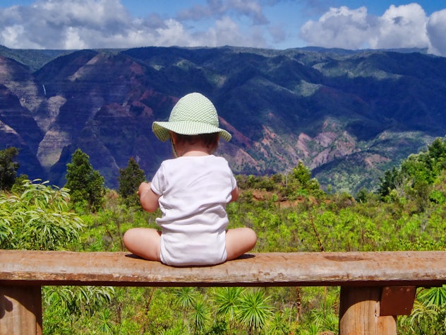 Baby wearing a sun hat facing lush, tropical mountain landscape