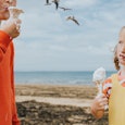 Comical image of two children standing on a sandy beach by the ocean. They both grip wafer cones con...
