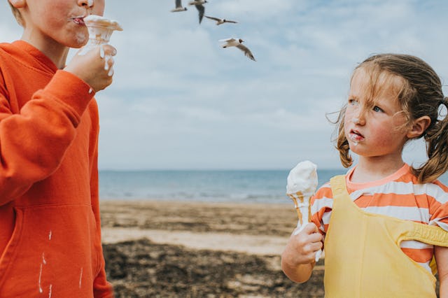 Comical image of two children standing on a sandy beach by the ocean. They both grip wafer cones con...