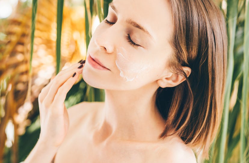 Smiling young woman applying moisturizer on face.