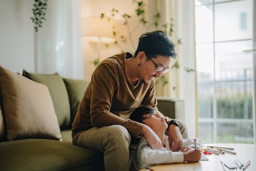 A dad sitting on a couch has his hands on his son's shoulders, as his son sits on the floor and colo...