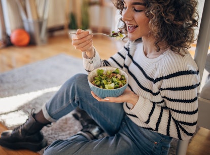 Young woman sitting on the floor at home and eating a salad.