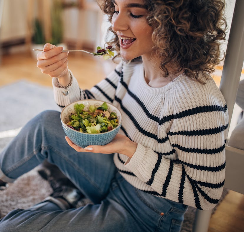 Young woman sitting on the floor at home and eating a salad.