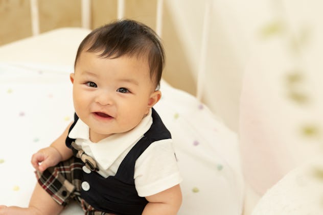 A baby wearing a bow tie and vest sitting in a crib smiling at camera, tudor baby names