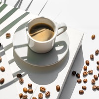 Aromatic morning coffee in a white porcelain cup and Roasted Coffee Beans with shadow from green pal...