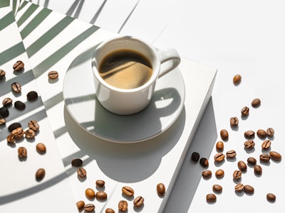 Aromatic morning coffee in a white porcelain cup and Roasted Coffee Beans with shadow from green pal...