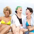 Three women laugh together as they enjoy a beach day and prepare for the 2022 summer solstice, which...