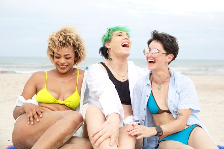 Three women laugh together as they enjoy a beach day and prepare for the 2022 summer solstice, which...