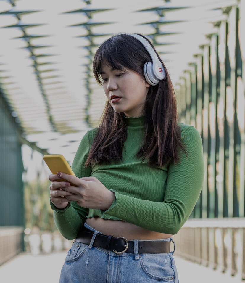 Side shot of young woman listening to audiobook through wireless headphones, blurred view of urban a...
