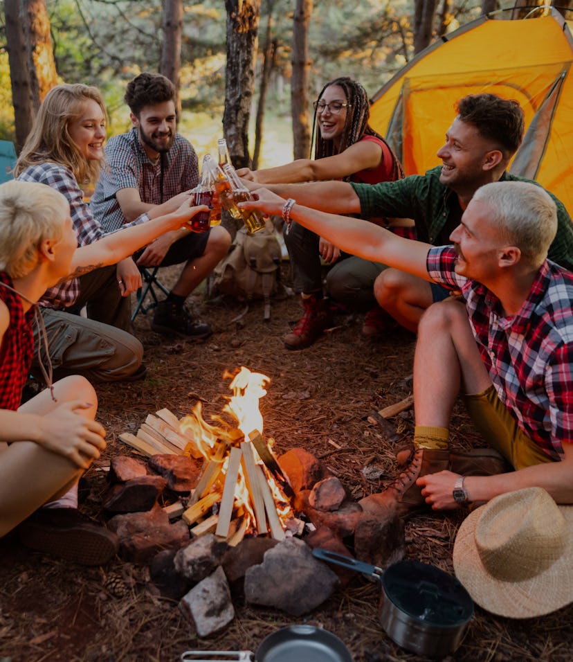 Group of young Caucasian friends sitting next to the bonfire and toasting with beer bottles in the w...