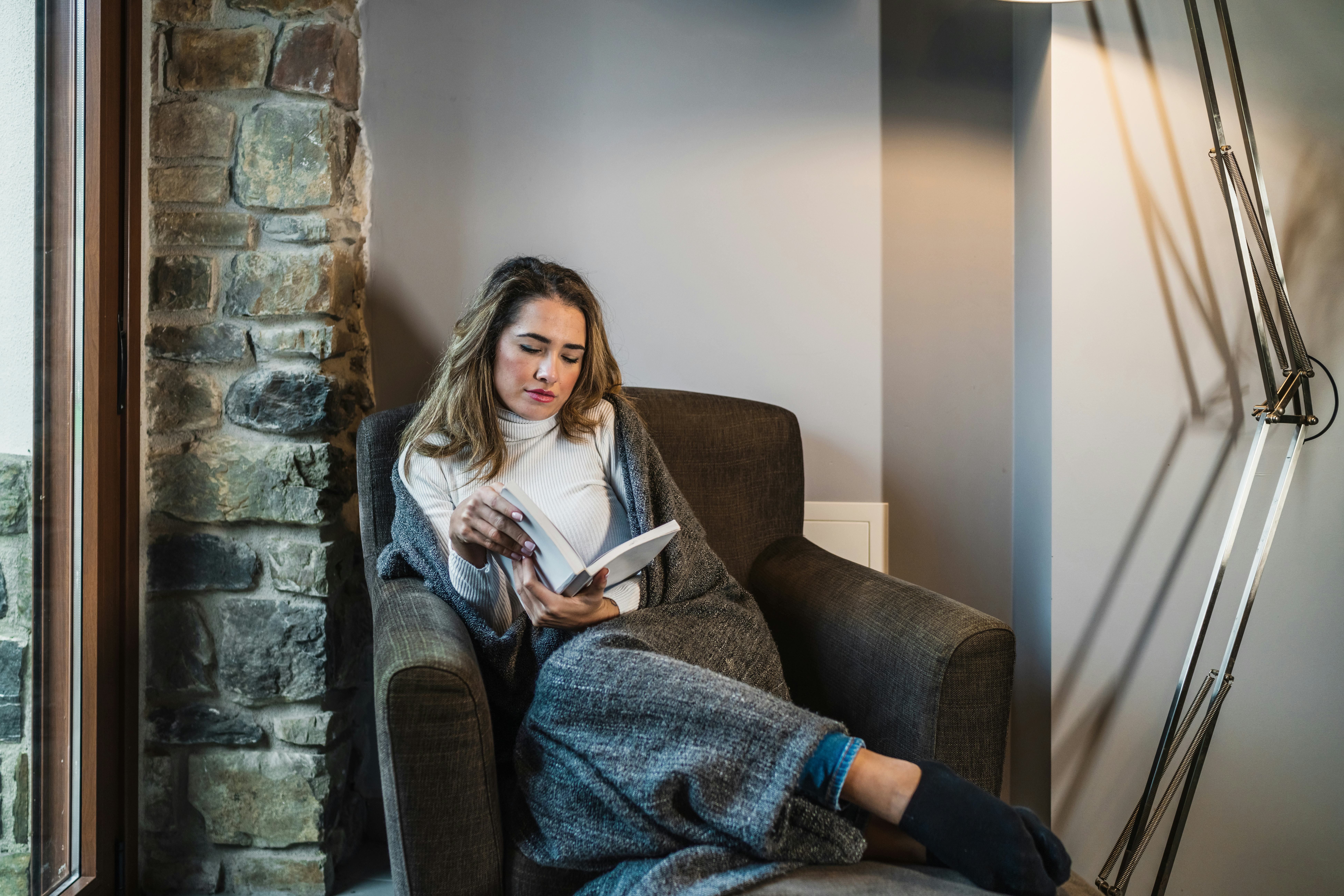 Close-up of a young blonde Caucasian woman reading a book in the warmth of her home on the sofa by t...
