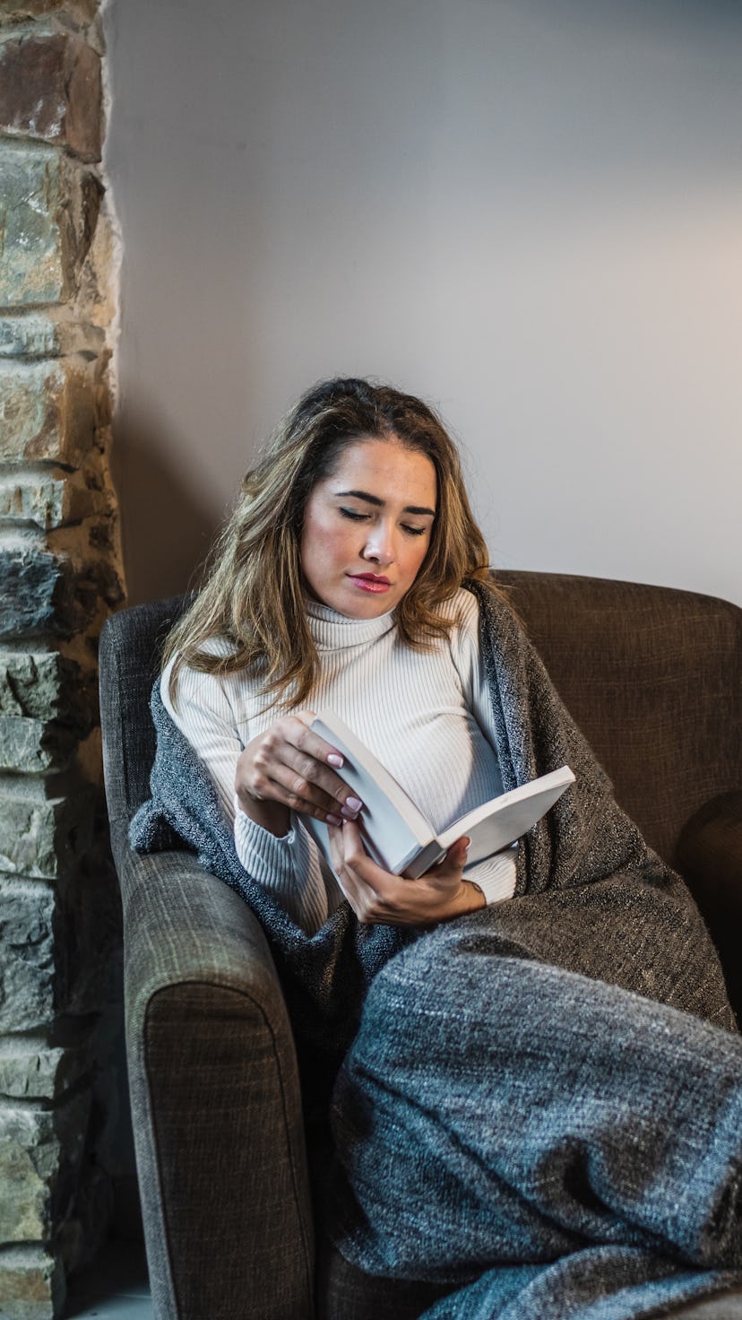 Close-up of a young blonde Caucasian woman reading a book in the warmth of her home on the sofa by t...