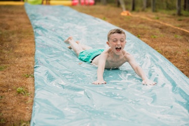 A front view shot of a young boy sliding down a water slide on his stomach in a garden, he is having...