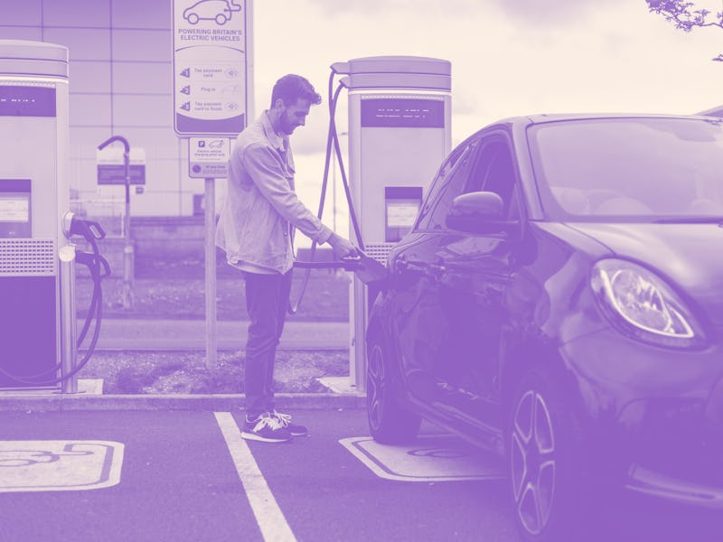 A young adult plugging a charger into his black car in a car park. He is smiling wearing casual clot...