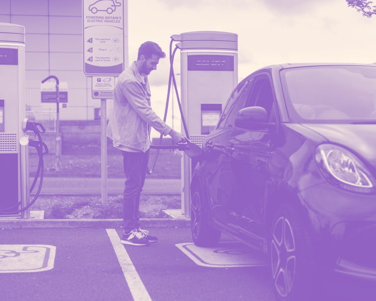 A young adult plugging a charger into his black car in a car park. He is smiling wearing casual clot...