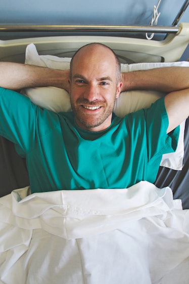 A friendly and bald Caucasian man with a beard lying with his hands behind his head smiling in a hos...