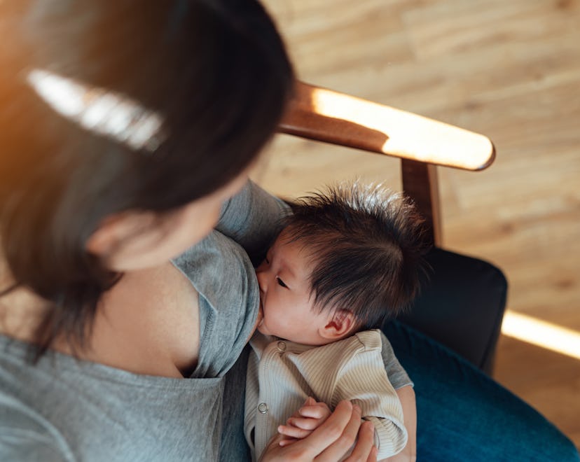 Over the shoulder view of mom breastfeeding newborn baby while sitting on armchair at home with war...