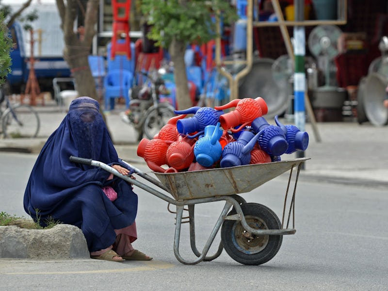 An Afghan burqa-clad woman sits next to a wheelbarrow loaded with plastic water cans along a street ...