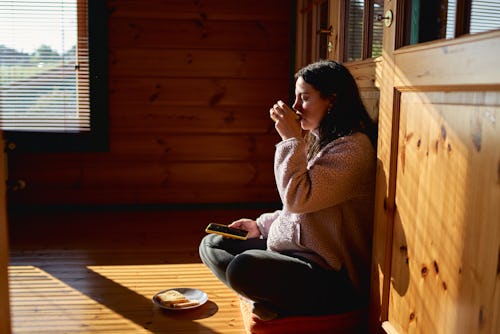 side view of a woman sitting in the sun while drinking coffee and toast and holding a cell phone in ...