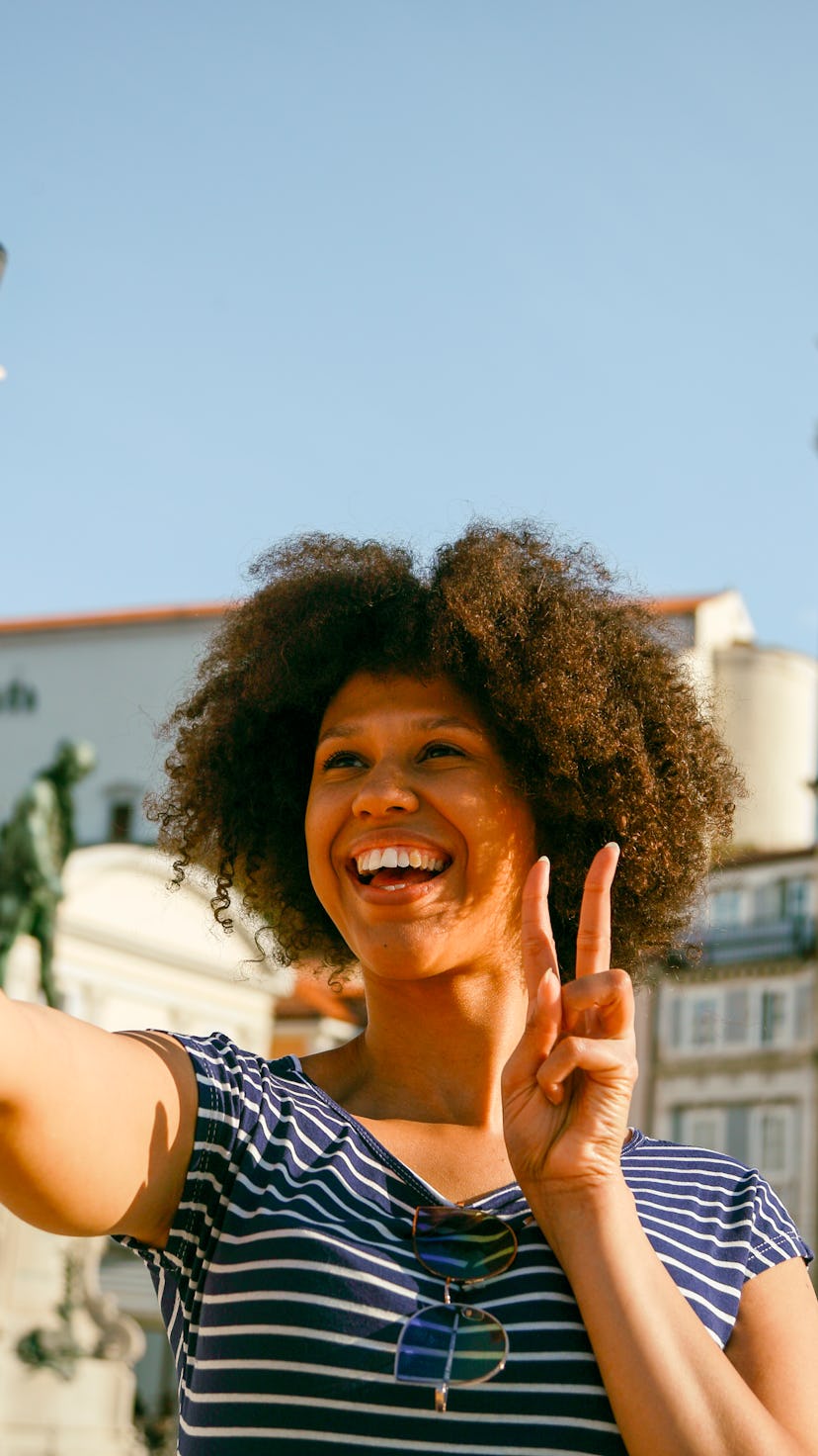Young woman holding two fingers for victory and taking a selfie in an old town center. Leo season is...
