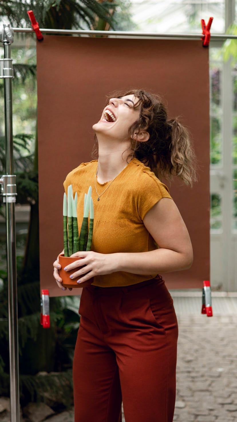 Cheerful woman standing in a botanical garden with a brown colour backdrop setup behind her holding ...