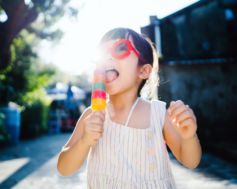 A girl with dark brown hair wearing red sunglasses and a dress licking a rainbow popsicle outside fo...