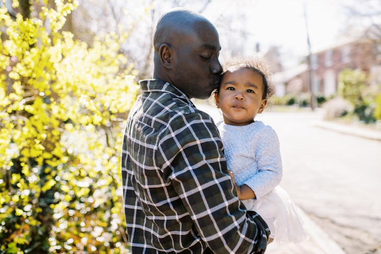 Father holding his small child in his arms and kissing their head while standing on the street on a ...