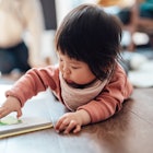 Cute Asian baby girl turning the page while reading a book, lying down on the floor at home. Childre...