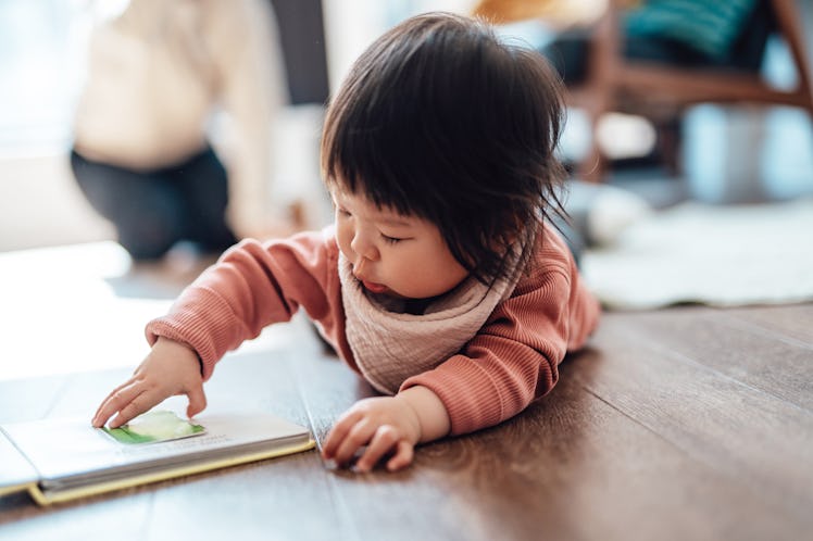 Cute Asian baby girl turning the page while reading a book, lying down on the floor at home. Childre...