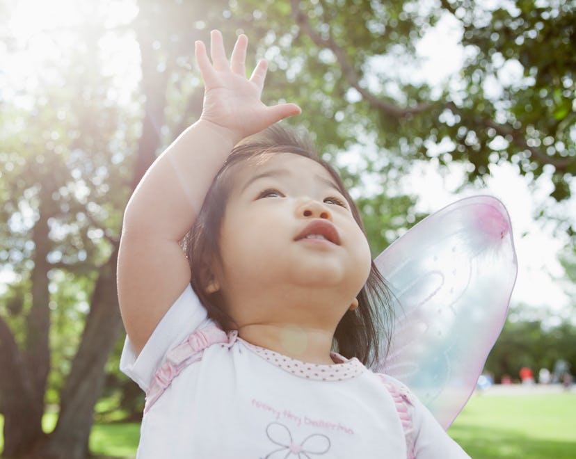 Toddler girl with dark hair wearing butterfly wings reaching up with an open palm, outside, in an ar...