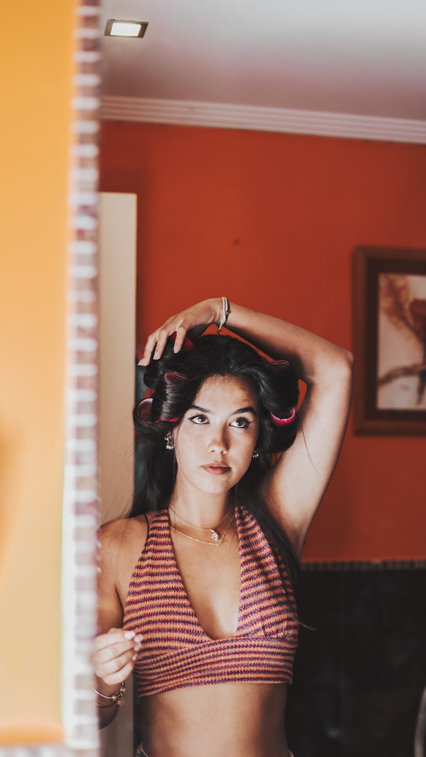 Young woman is posing in front of a mirror, staring at her reflection with curlers in her hair. The ...