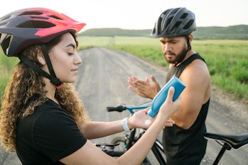 A young latin couple on their bikes, putting on lotion before cycling on a road outside.